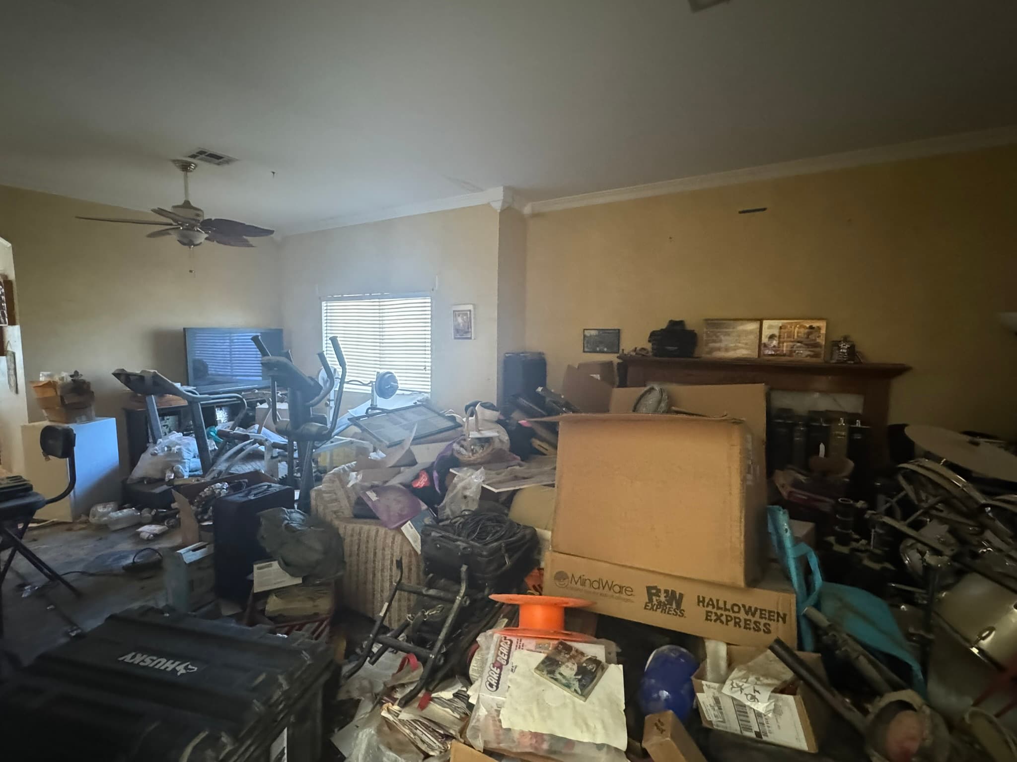 Cluttered living room packed with boxes, exercise equipment, and belongings piled across the floor